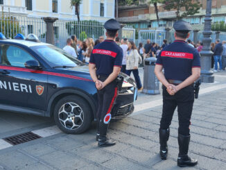 Carabinieri in divisa davanti all'ingresso di un edificio scolastico, con studenti e passanti sullo sfondo.