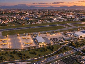 Vista notturna dell'aeroporto di Capodichino con pista e terminal illuminati, con la città sullo sfondo.