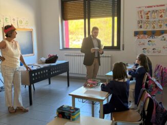 Classe scolastica con studenti seduti ai banchi e insegnanti o amministratori in piedi in aula durante il primo giorno di scuola.