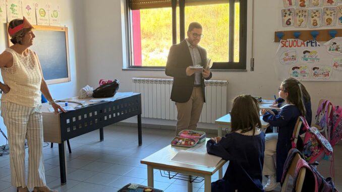 Classe scolastica con studenti seduti ai banchi e insegnanti o amministratori in piedi in aula durante il primo giorno di scuola.