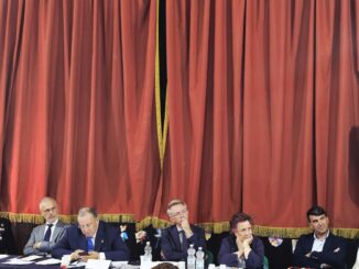 Incontro pubblico in una sala con persone sedute di fronte a un palco con tavolo e microfoni, decorazioni colorate e un grande tendone rosso sullo sfondo.